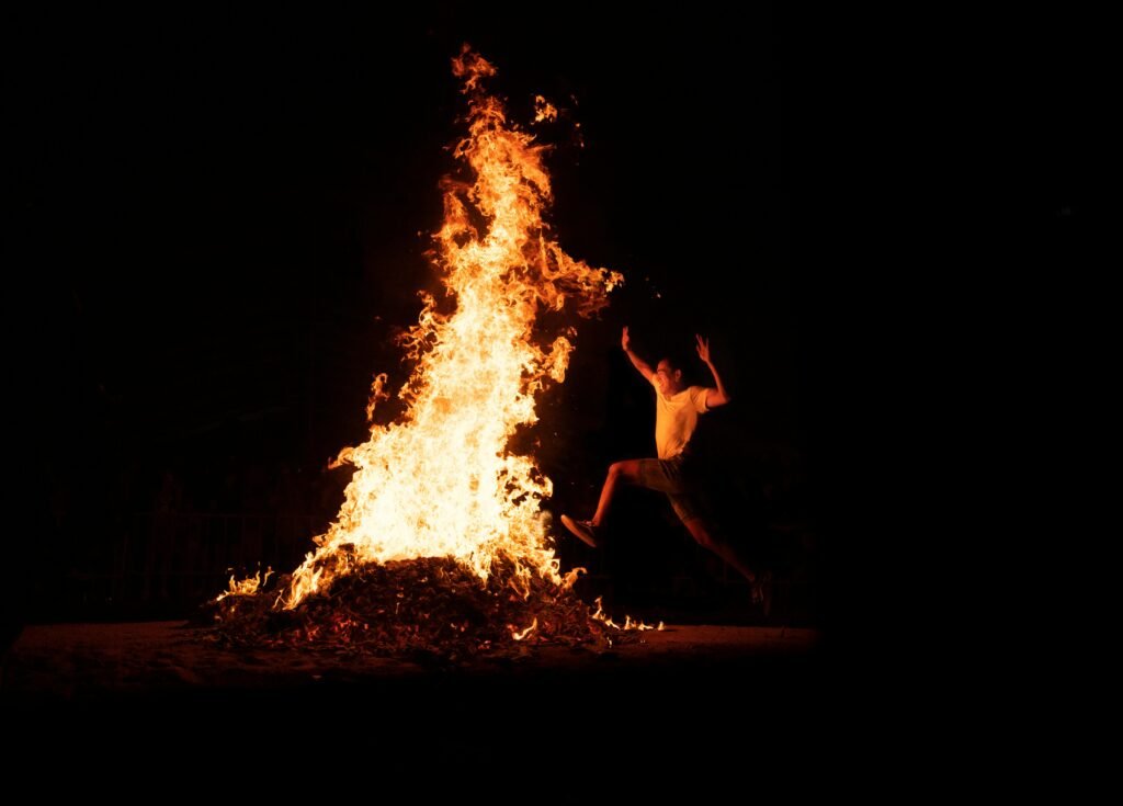 An adventurous man leaps over a large bonfire during a night celebration, highlighting energy and tradition.